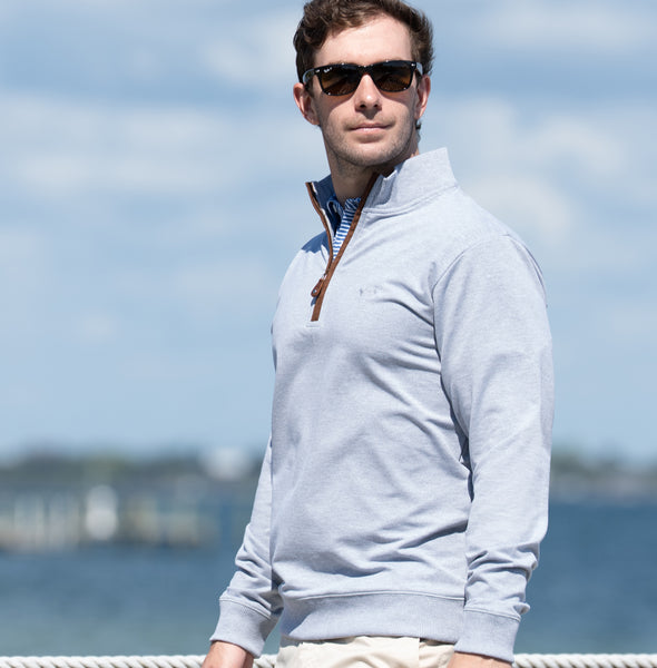 Man wearing a light gray pullover with a zipper, sunglasses, and beige pants standing by a body of water.