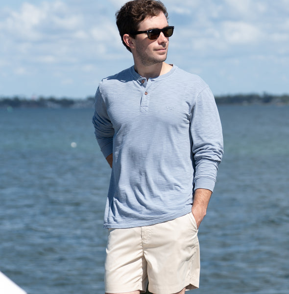 Man wearing sunglasses and a light blue long-sleeve shirt with beige shorts by a body of water.